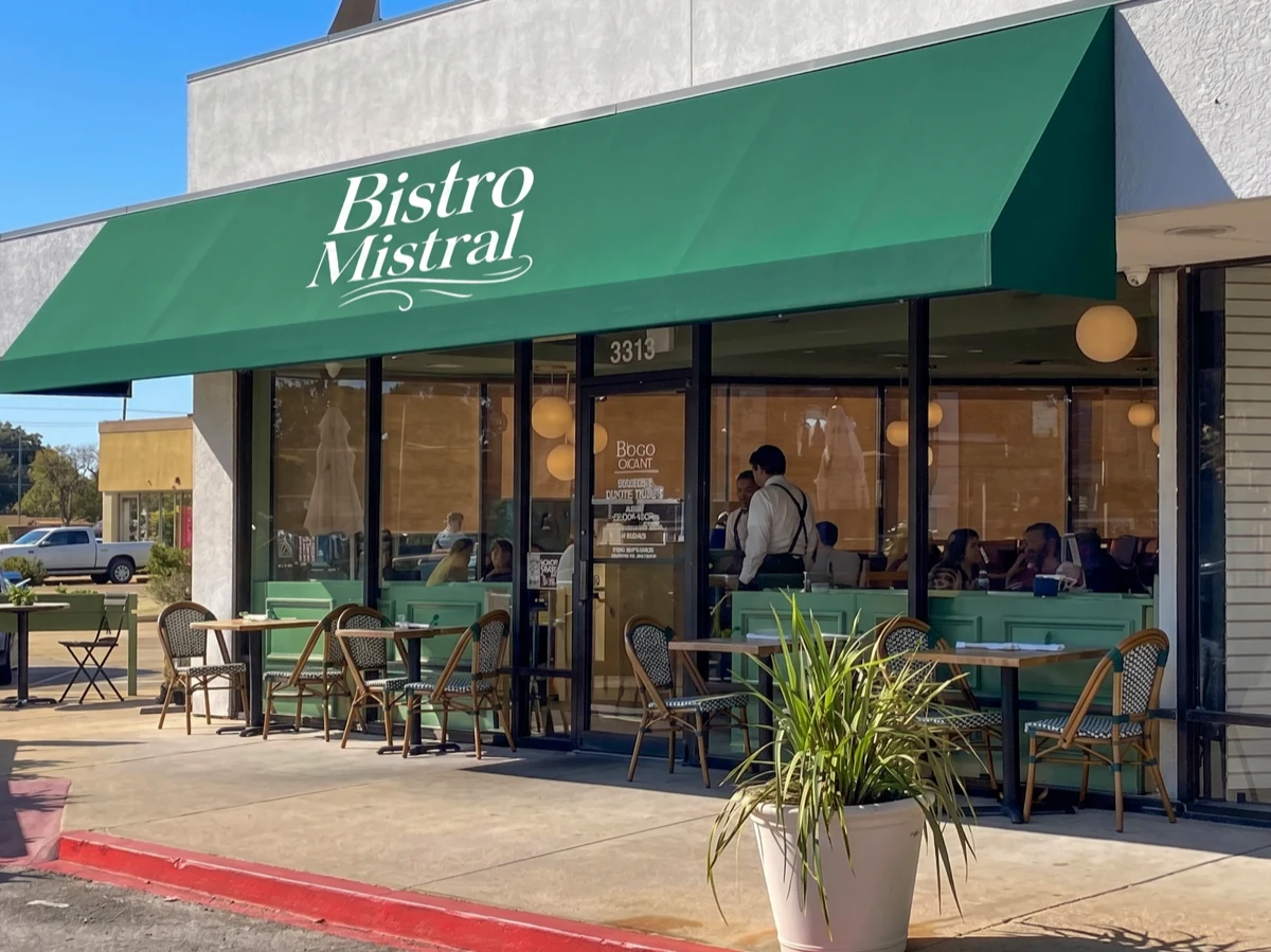 Bistro Mistral Bellaire facade — sage green awning with script signage, warm dining room visible through picture windows, and a Parisian-style patio with cane bistro chairs